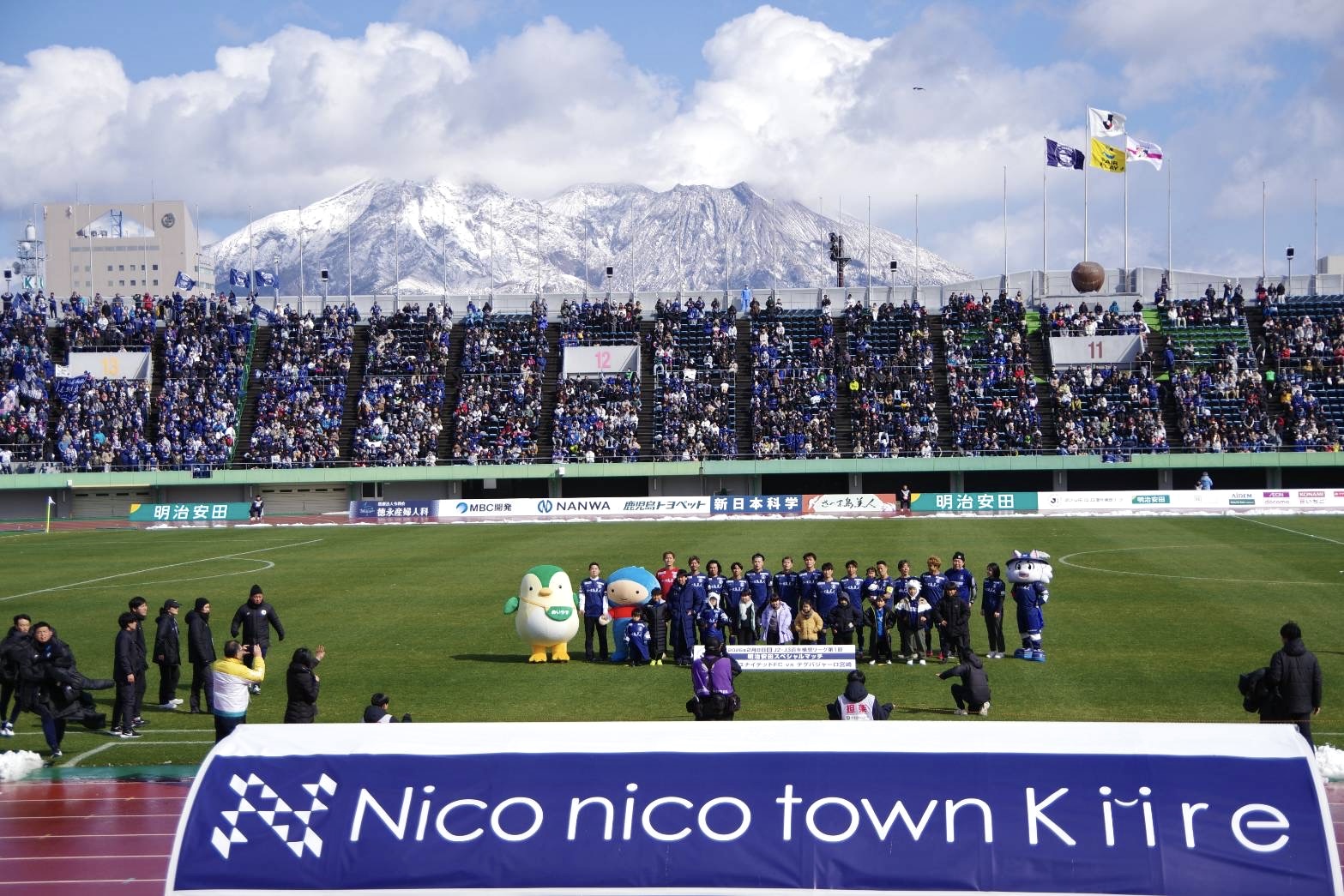 雪化粧の桜島を背景に、白波スタジアムのピッチで鹿児島ユナイテッドFCの選手たちが試合前に記念撮影をしている様子。手前には「Nico nico town Kiire」のロゴ入りバナーが写っている。