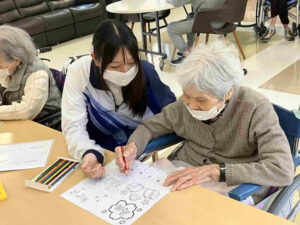 住宅型有料老人ホームニコニコハウスで体験学習中の学生が職員とともに利用者様と交流しながら塗り絵を行っている様子