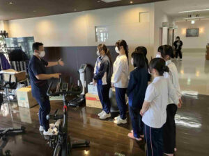 高校生がデイケア施設でスタッフからリハビリ機器の使い方について説明を受けている様子