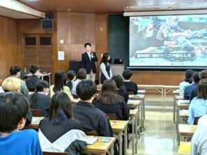 鹿児島国際大学の教室で学生約60名に対し、スクリーンを使って介護の魅力や現場について説明する卒業生スタッフ