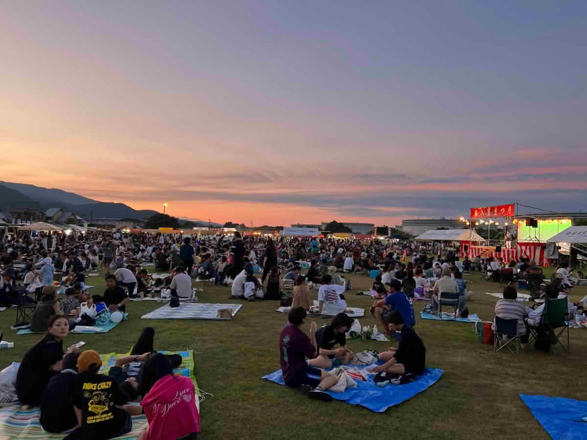 夕焼け空の下、マリンピア喜入の芝生広場に多くの来場者が集まり、ステージや屋台を楽しむ第39回喜入夏まつりの様子