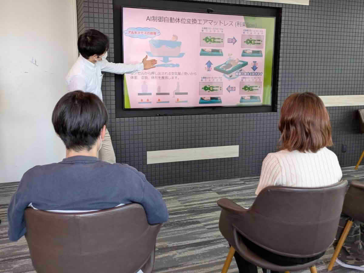 スクリーンにAI制御自動体位変換エアマットレスの資料を映し、学生へ仕組みを説明する職員
