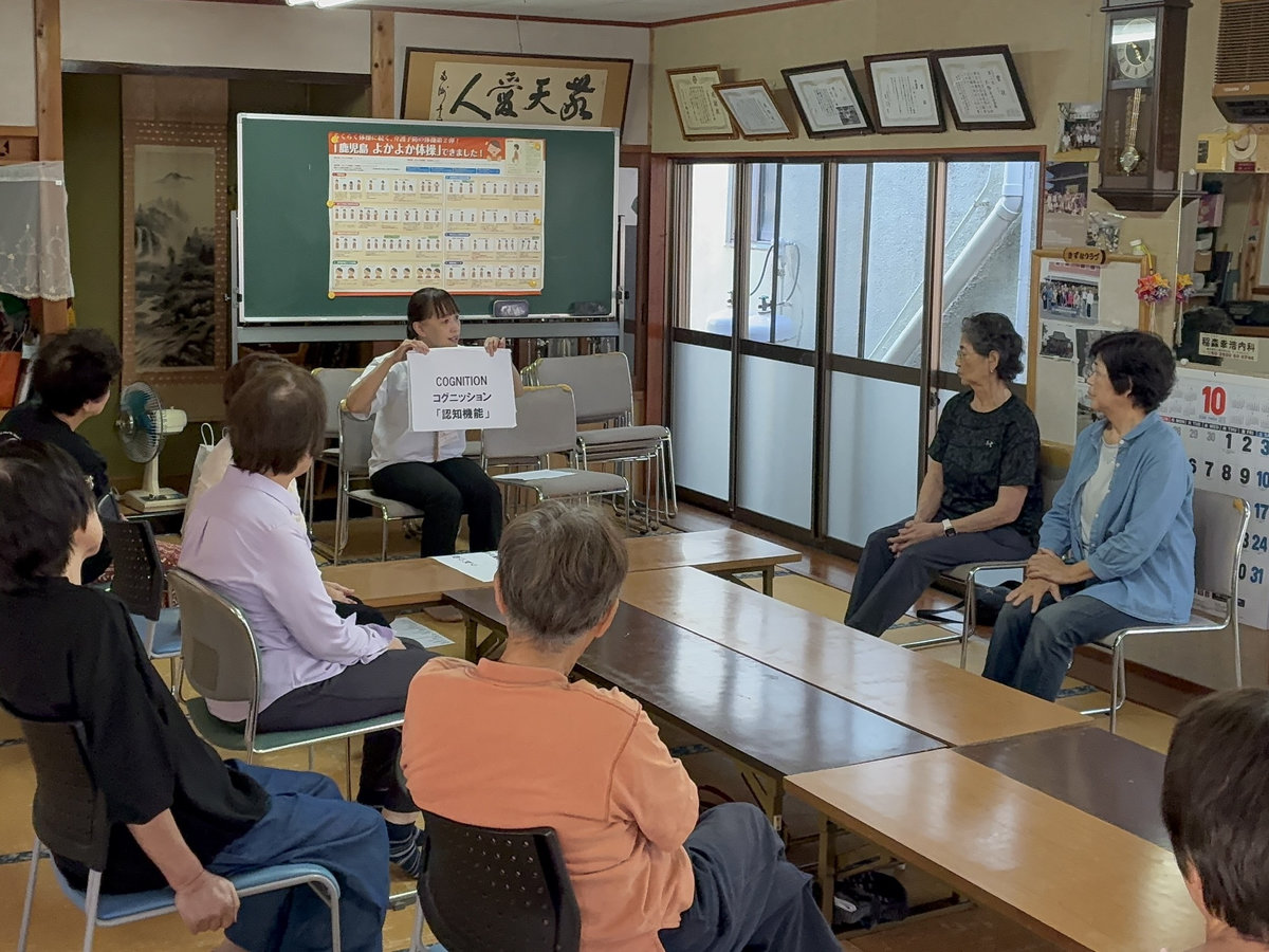 鹿児島市西谷山の不動寺公民館で、講師が「COGNITION（認知機能）」と書かれたカードを示し、コグニサイズについて説明する様子