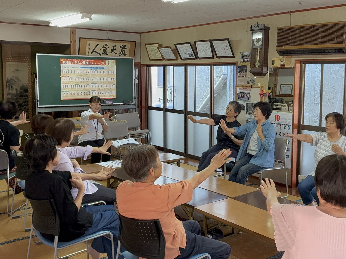 鹿児島市西谷山の不動寺公民館で、講師の動きに合わせて参加者が椅子に座り、腕を伸ばす運動に取り組む様子