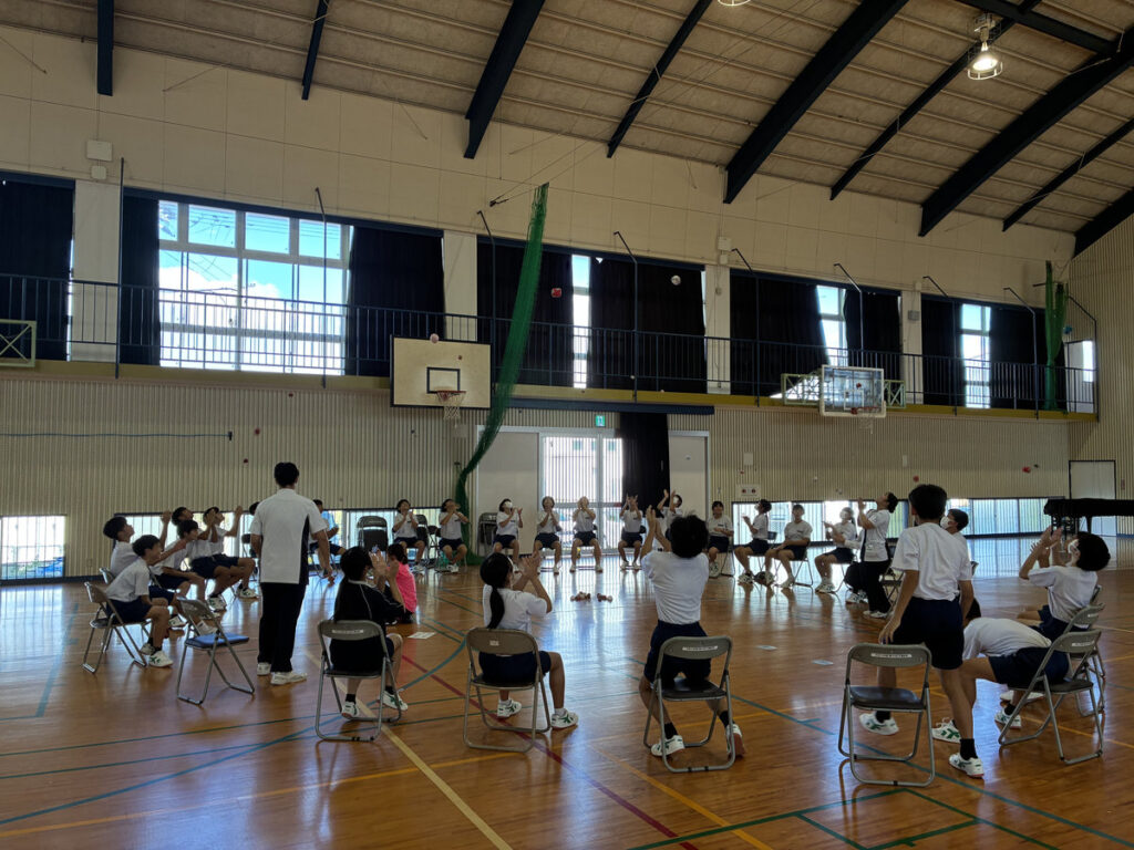 南指宿中学校の体育館で、生徒たちが円形に並んだ椅子に座り、スタッフと一緒にコグニサイズを体験している様子